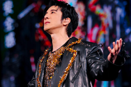 Wang Leehom performing on stage in an ornate black leather jacket with gold embellishments, looking upward with colorful stage lights